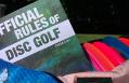 A picture of the most recent print edition of the Official Rules of Disc Golf and Competition Manual for Disc Golf Events. The book is sitting in a pink diamond ripstop duffel bag next to a blue towel and an array of red and blue discs.
